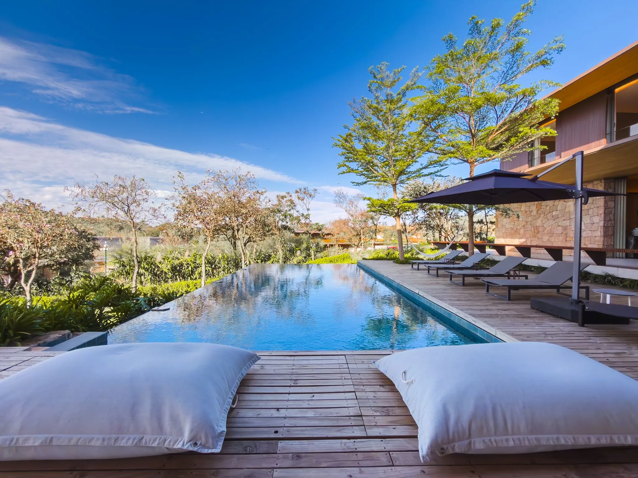 Deck com almofadas diante de uma piscina longa, com árvores e céu azul