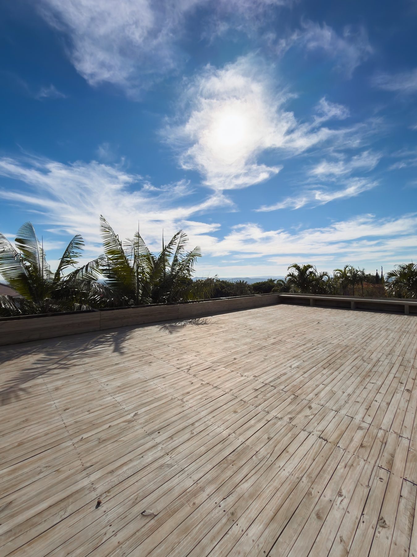Deck de madeira do rooftop sob o sol, com palmeiras e o horizonte ao fundo