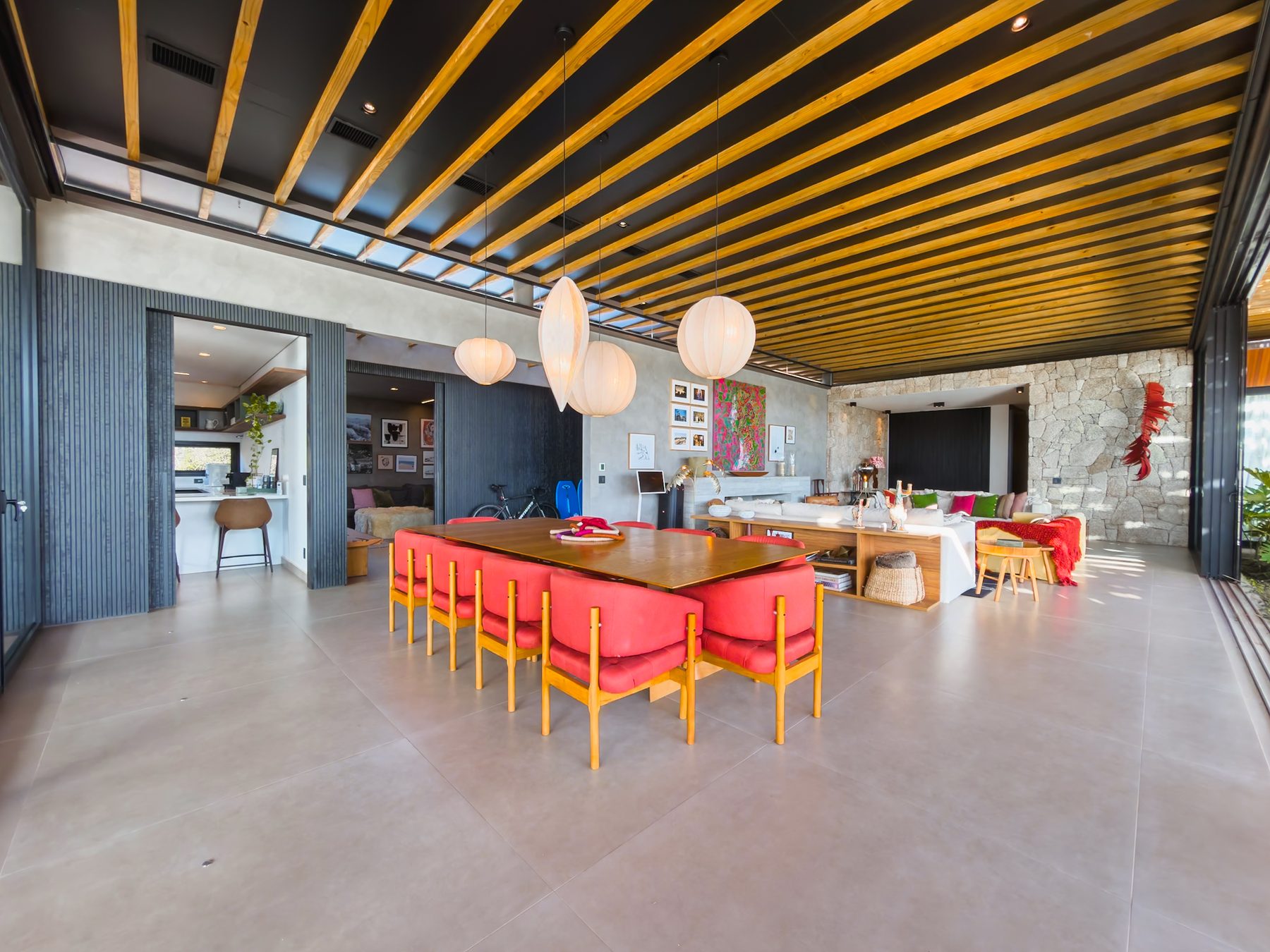 Sala de jantar com mesa longa de madeira, cadeiras vermelhas e luminárias de papel, aberta para o jardim