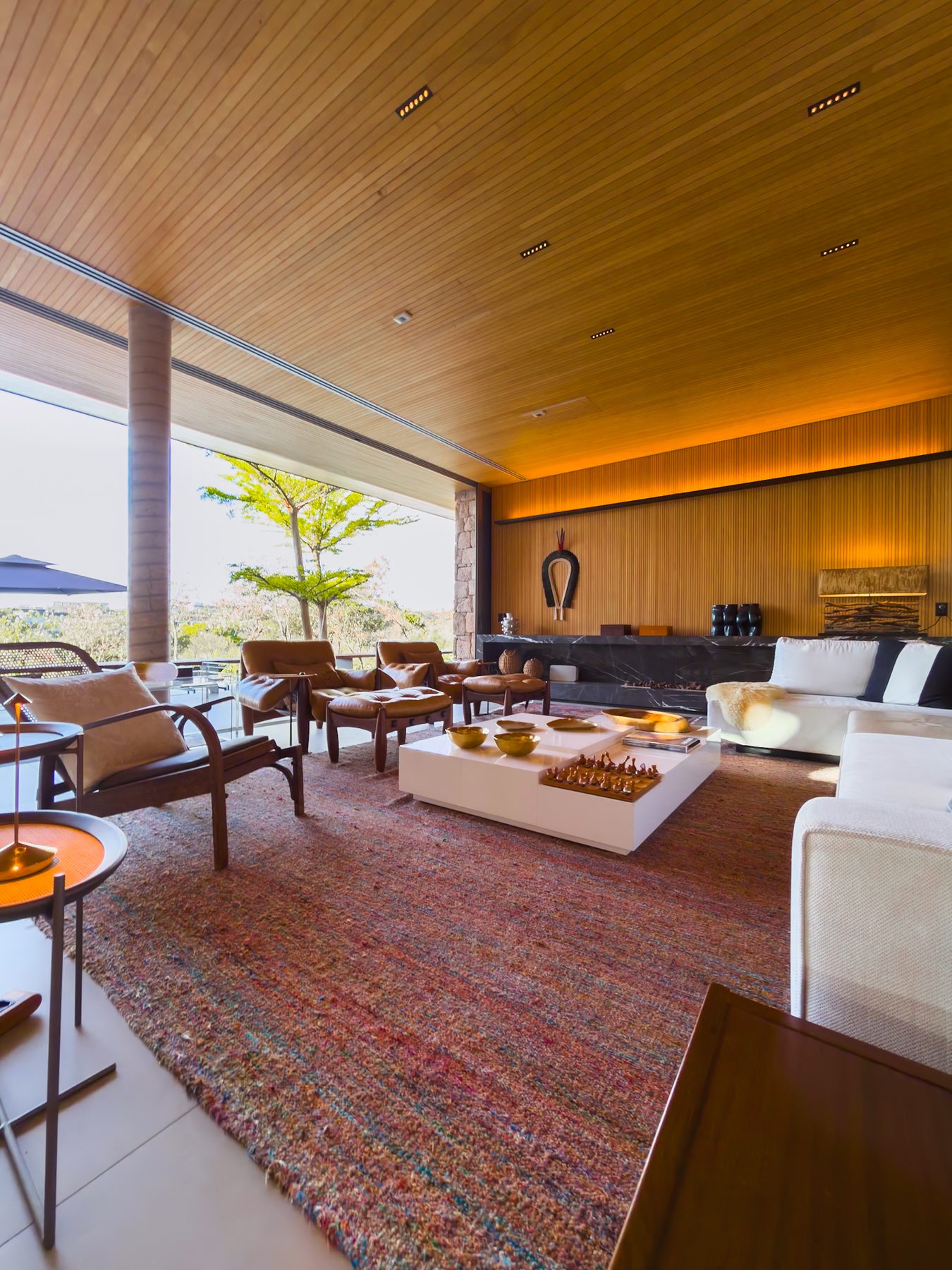 Living amplo sob forro de madeira, com tapete vermelho, poltronas de couro e vista para o jardim
