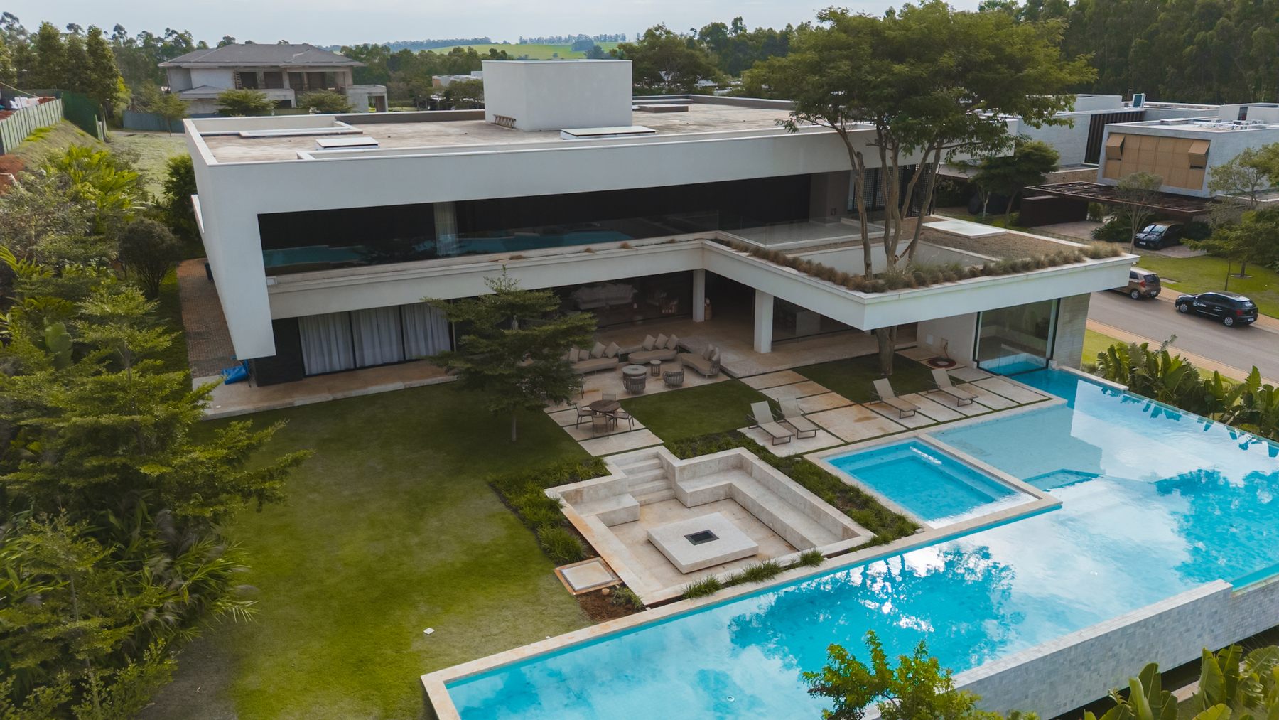 Vista aérea da casa branca de dois pavimentos, com a piscina em L, a praça de fogo e o jardim
