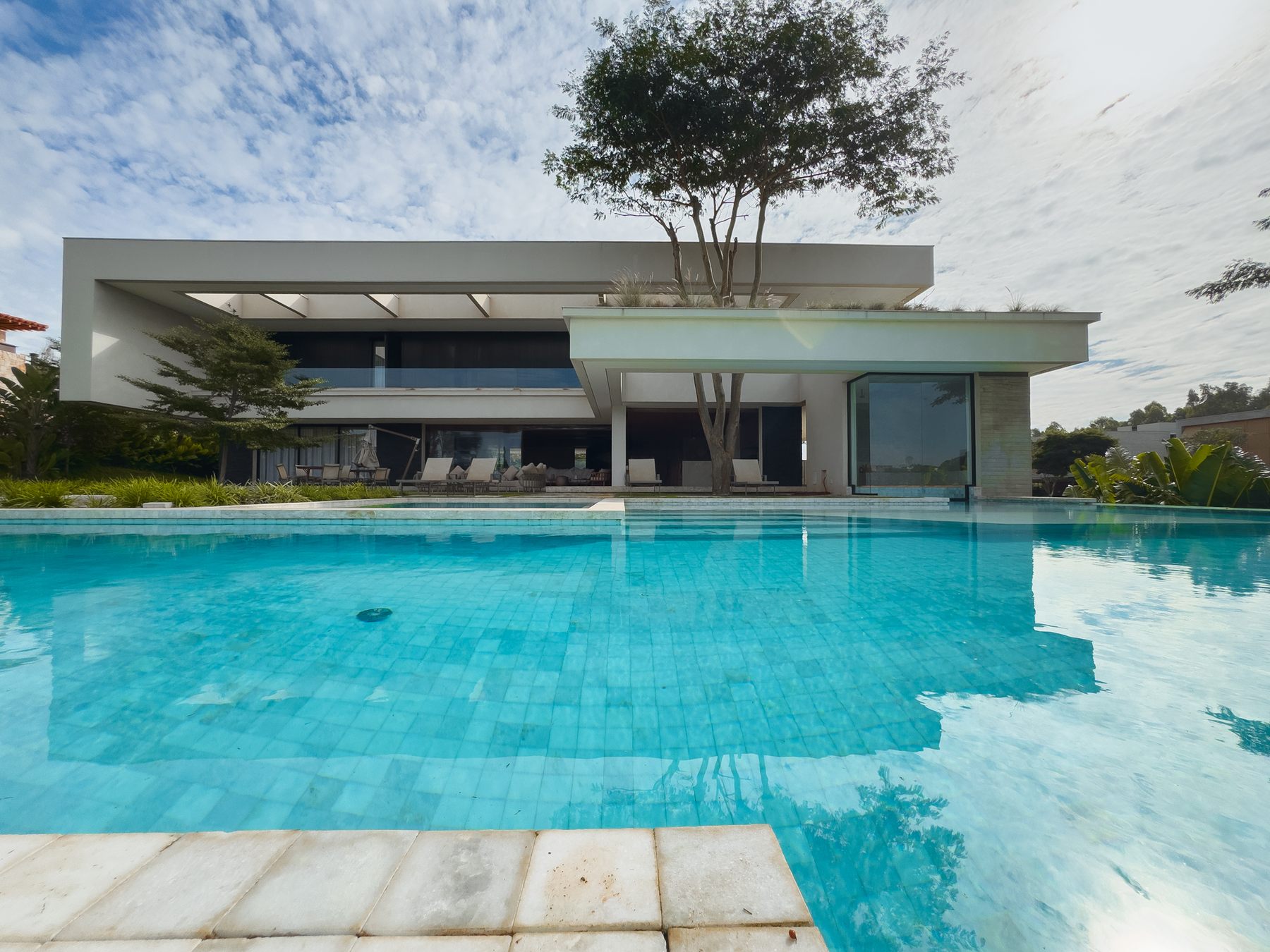 Piscina diante da casa branca de dois pavimentos, com uma árvore sobre o terraço e céu com nuvens