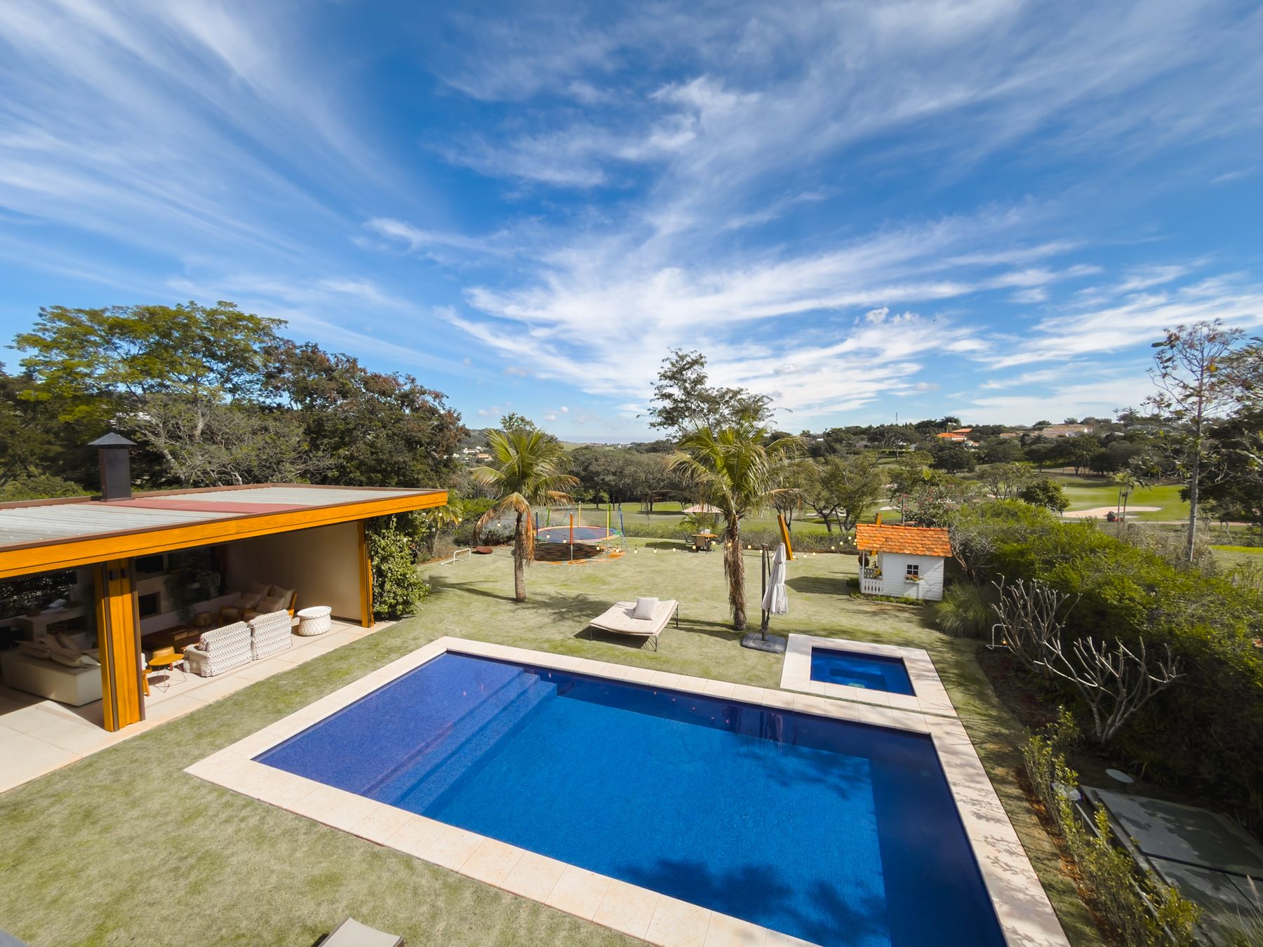 A piscina e o pergolado da área gourmet vistos do alto, com o gramado e o campo de golfe ao fundo, sob céu com nuvens