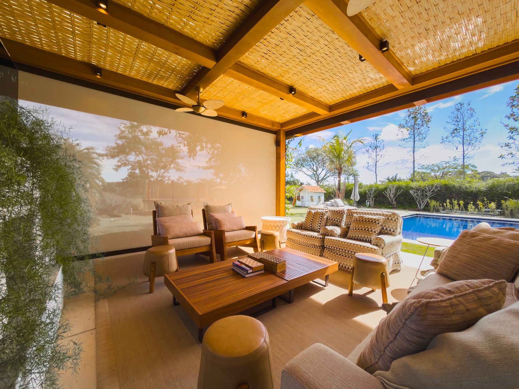Sofás e mesa de centro de madeira sob o pergolado, com a piscina e o jardim ao fundo