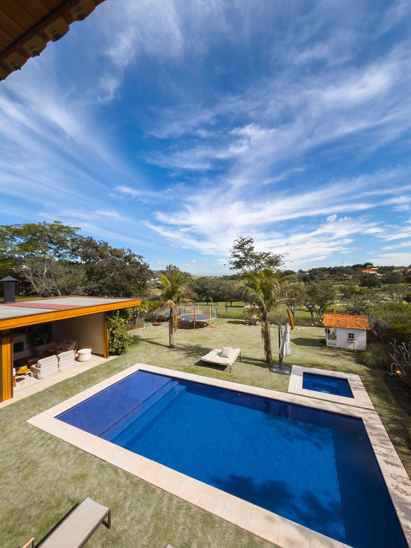 A piscina vista do pavimento superior, com o pergolado da área gourmet, o gramado e o céu com nuvens