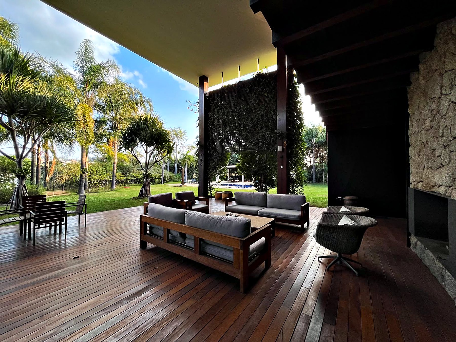 Deck de madeira coberto com sofás, poltronas e a lareira na parede de pedra, aberto para o jardim